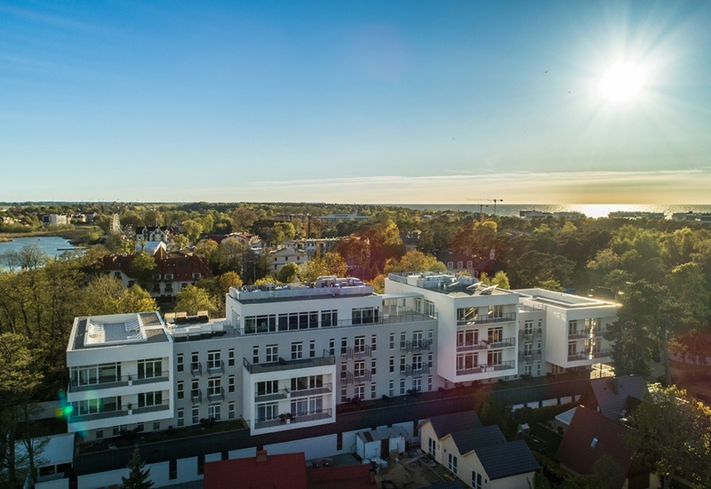 Nowoczesny biały hotel przy wybrzeżu z balkonami i tarasem na dachu, widok na zalesnioną lagunę i błyszczące morze w jasnym popołudniowym słońcu.