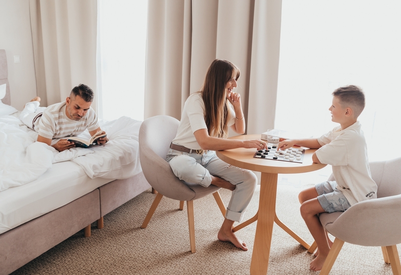 Jasny, rodzinny pokój hotelowy w stonowanych barwach z dużym oknem; ojciec czyta na łóżku, mama z synem grają w warcaby przy stoliku.