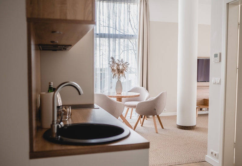 Przytulny apartament z aneksem kuchennym, okrągłym stolikiem z suszonymi kwiatami, trzema wygodnymi fotelami i dużym oknem z firaną.