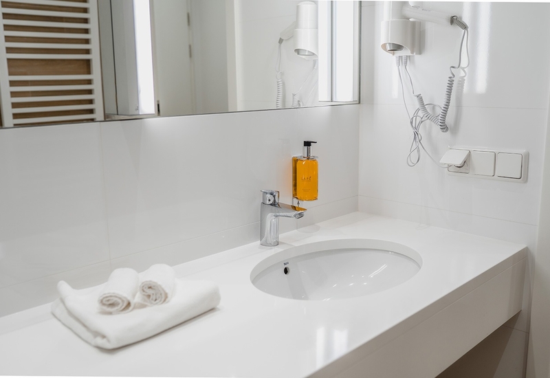 Jasna nowoczesna łazienka hotelowa z owalną umywalką, chromowaną baterią, suszarką ścienną, dozownikiem mydła i złożonymi białymi ręcznikami.