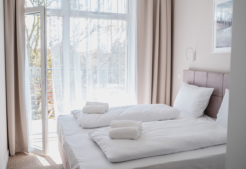 Jasny pokój z dwoma łóżkami, białą pościelą i zrolowanymi ręcznikami, różowym tapicerowanym zagłówkiem i drzwiami na balkon z lekkimi firankami.