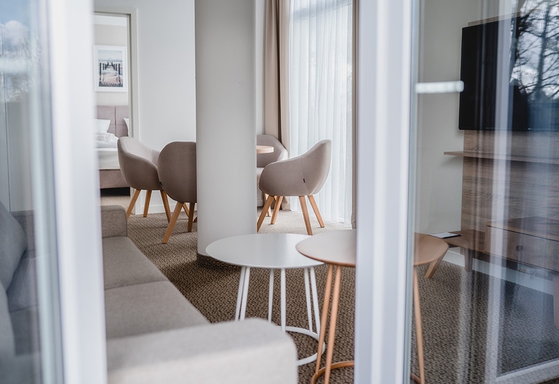 Nowoczesny apartament hotelowy z salonem: neutralne fotele przy małym stoliku, sofa, telewizor i firany
