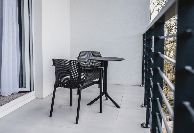 Balkon przy pokoju z dwoma czarnymi krzesłami z siatki i małym okrągłym stolikiem, przesuwnymi drzwiami z lekką firaną oraz metalową balustradą.