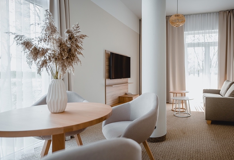 Jasna, nowoczesna część dzienna apartamentu z okrągłym drewnianym stołem i wazonem suchych traw, dwoma fotelami, sofą, telewizorem i zwiewnymi firankami.