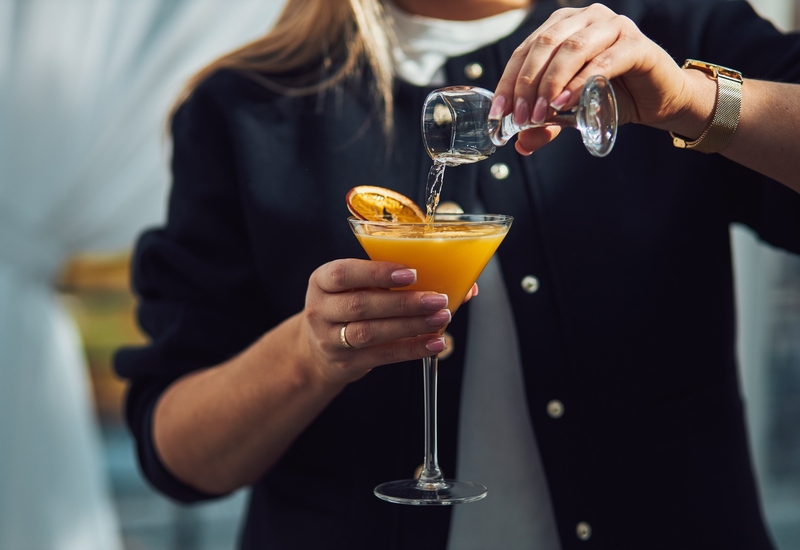 Barman nalewa klarowny alkohol do intensywnie pomarańczowego koktajlu ozdobionego suszoną plasterką pomarańczy w kieliszku