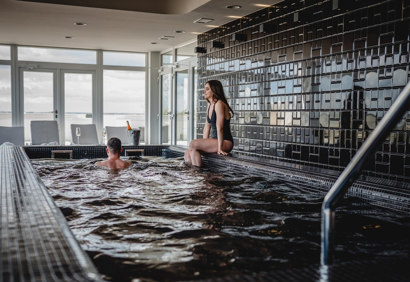 Wewnętrzne jacuzzi hotelowe z błyszczącymi czarnymi płytkami, kobieta siedzi na krawędzi, gość w wodzie, szampan w wiaderku i widok na morze.