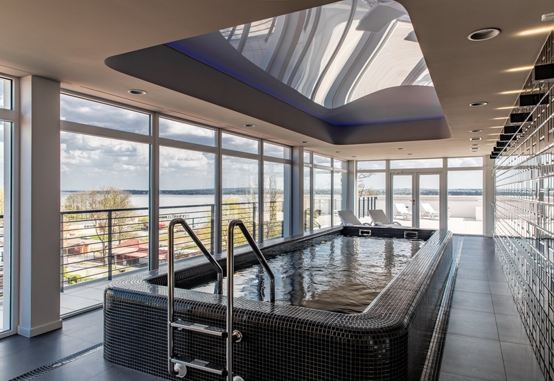Wewnętrzne spa z czarnym, mozaikowym basenem, stalową drabinką, panoramicznymi oknami z widokiem na jezioro i leżakami