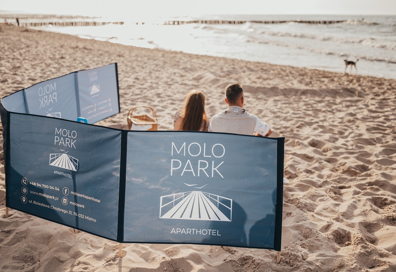 Para siedzi za parawanem z logo Molo Park na piaszczystej plaży, koszyk piknikowy obok, widok na morze o złotej godzinie