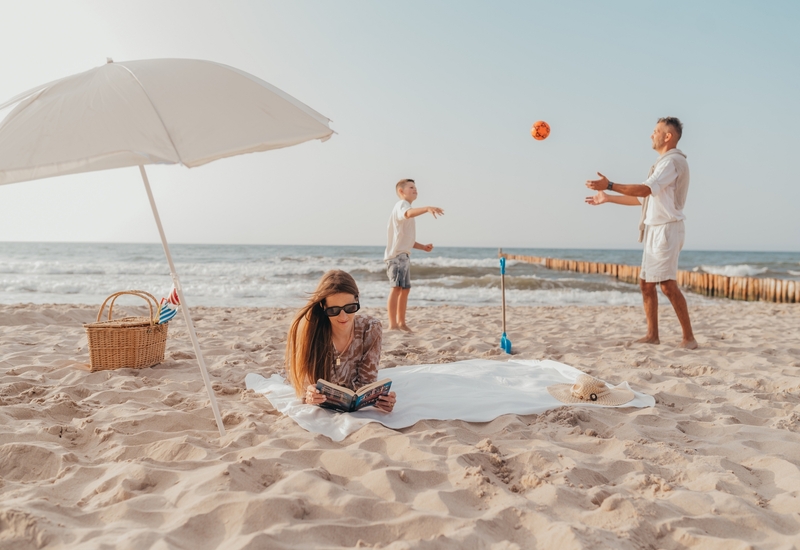 Rodzinna chwila na plaży: kobieta czyta pod parasolem, koszyk piknikowy i kapelusz obok; ojciec z synem rzucają piłkę.