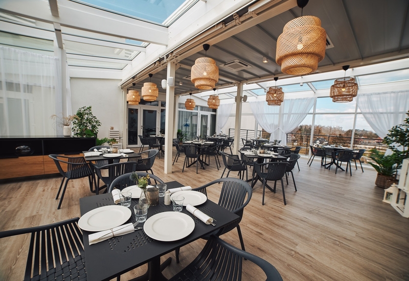 Jadalnia hotelowa z przeszklonym dachem, rattanowymi lampami wiszącymi, nakrytymi czarnymi stołami i krzesłami, lekkimi białymi zasłonami i panoramicznym widokiem