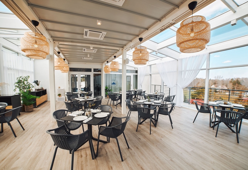 Jasna restauracja z przeszklonym dachem, wiklinowymi lampami, czarnymi krzesłami, nakrytymi stolikami i widokiem na drzewa