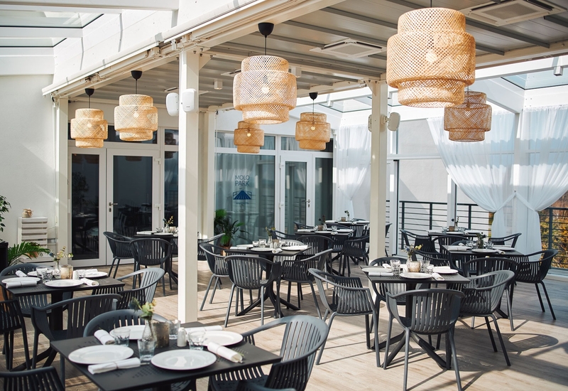 Kryty taras restauracyjny z plecionymi lampami rattanowymi, czarnymi stolikami i krzesłami nakrytymi do posiłków, zwiewne białe zasłony.