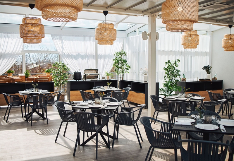 Jasna restauracja w oranżerii z wiklinowymi lampami, nakrytymi stolikami, nowoczesnymi czarnymi krzesłami i bufetem.
