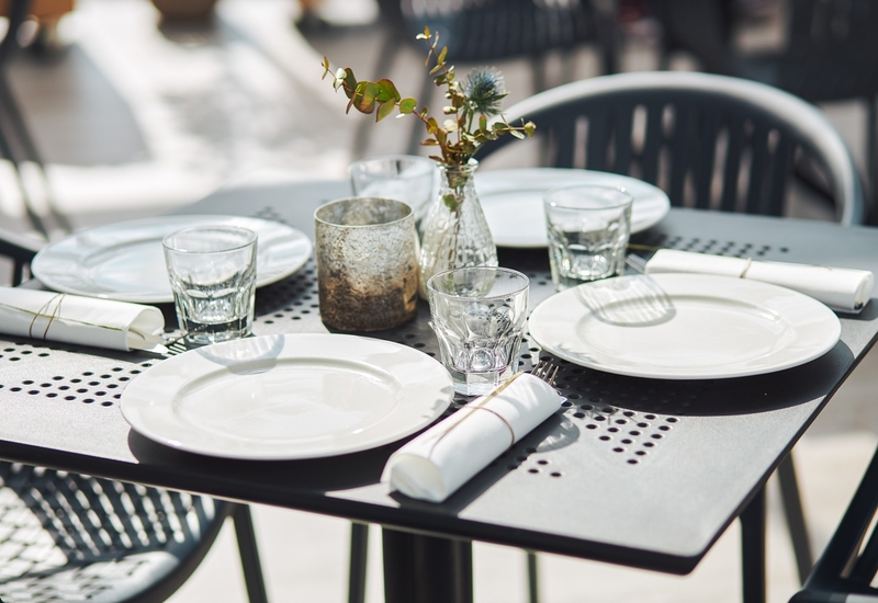 Zewnętrzny stolik restauracyjny z białymi talerzami, szklankami, zwiniętymi serwetkami i małym wazonem z zielenią.