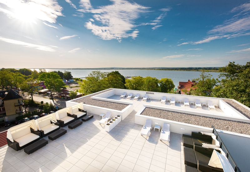 Taras na dachu z białymi leżakami, strefą lounge i miejscami do siedzenia, panoramiczny widok na spokojne jezioro i zadrzewiony brzeg