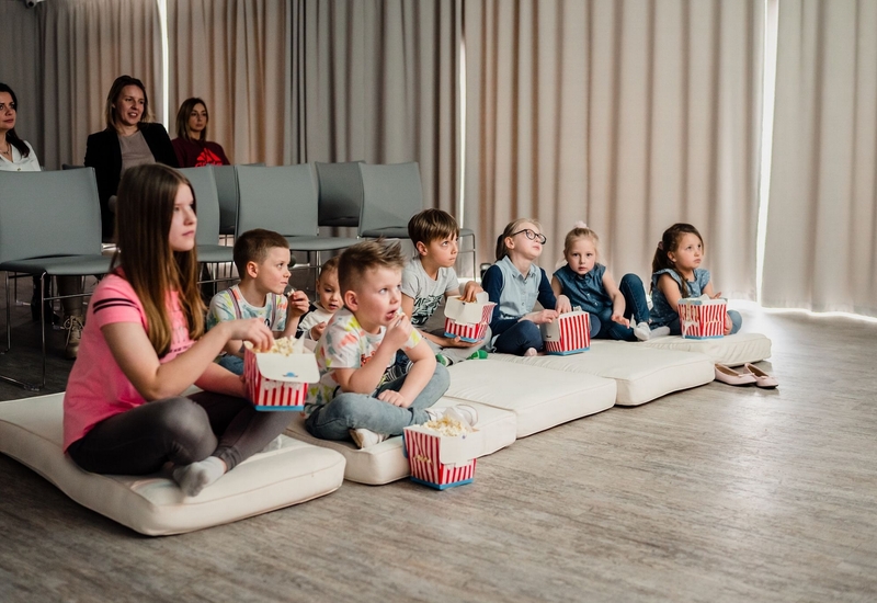 Dzieci na poduchach w sali hotelowej oglądają film i jedzą popcorn; dorośli siedzą na krzesłach za zasłonami.