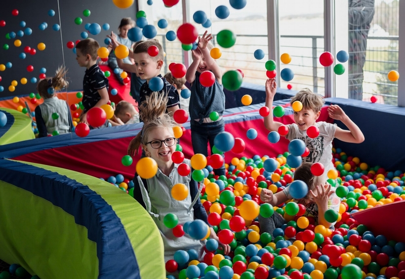 Rodzinne centrum zabaw z dużym kolorowym basenem kulkowym, miękkimi siedziskami i bawiącymi się dziećmi przy przeszklonej ścianie