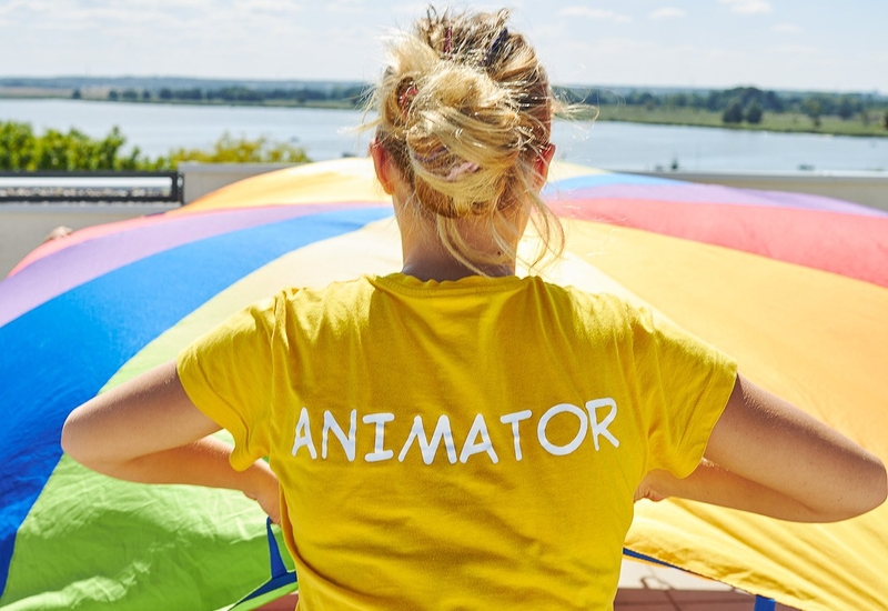 Animatorka w żółtej koszulce prowadzi zabawę z kolorową chustą na słonecznym tarasie hotelu z widokiem na jezioro