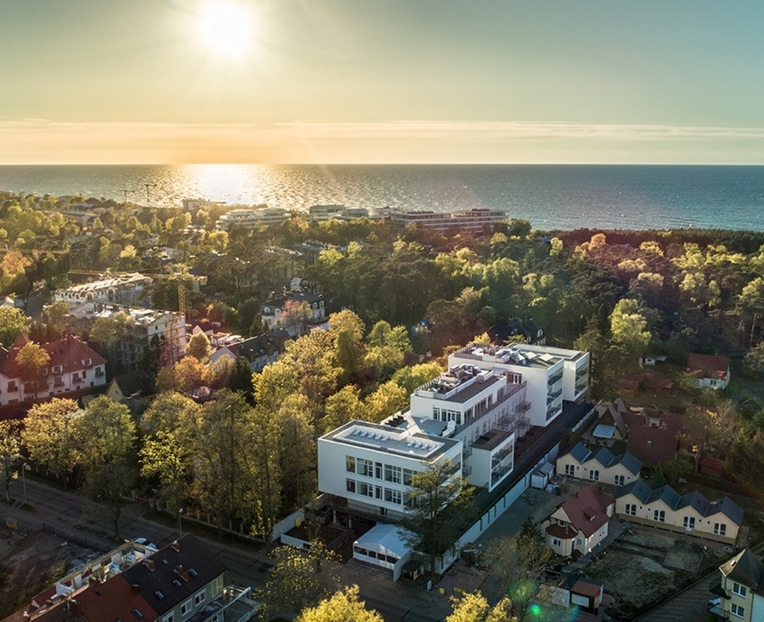 Nowoczesny biały hotel z tarasami i widokiem na morze, otoczony zielonymi drzewami i nadmorską promenadą o zachodzie słońca.