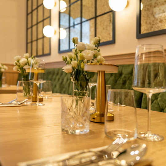Restauracja z drewnianym stołem, luksusową zieloną tapicerką i eleganckimi lampami, z kwiatami na każdym stole.