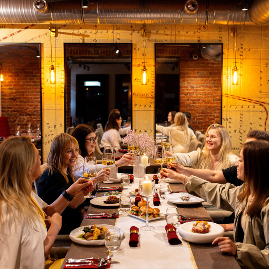 Grupa toastująca białym winem przy długim stole w przytulnej restauracji z odsłoniętą cegłą i ciepłym oświetleniem