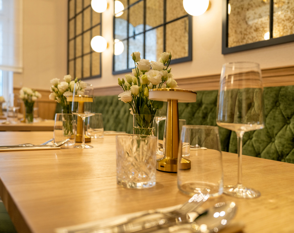 Restauracja z drewnianym stołem, luksusową zieloną tapicerką i eleganckimi lampami, z kwiatami na każdym stole.