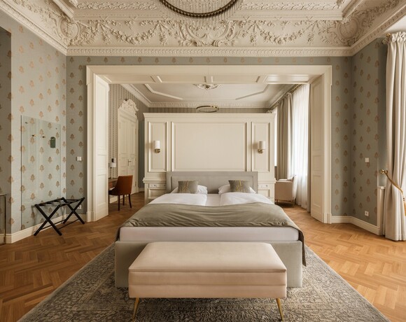 Luxuriöse Suite mit Kingsize‑Bett, opulenter Stuckdecke, Parkettboden, Sitz- und Schreibtischbereich und bodenlangen Vorhängen