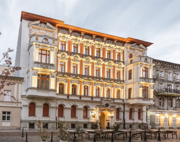 Ozdobna fasada hotelu z detalami sztukatorskimi, ciepłe oświetlenie, łukowe wejście i brukowany taras z stolikami.