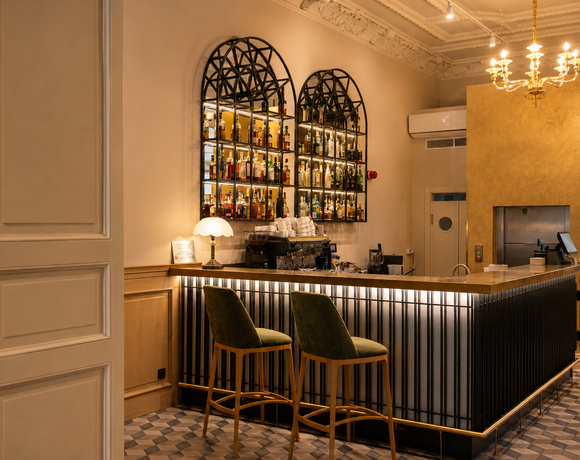 Intimate hotel bar with two green velvet stools, backlit arched liquor shelves, espresso machine, brass-accented counter and ornate ceiling chandelier