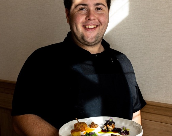 Uśmiechnięty kucharz w czarnym mundurze prezentuje białą paterę z finezyjnymi porcjami w przytulnej restauracji hotelowej.