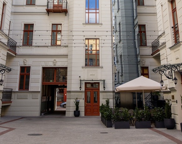 Zabytkowe białe podwórze z łukowymi oknami, szklaną windą, tarasem z ogródkiem pod parasolem i lampkami.
