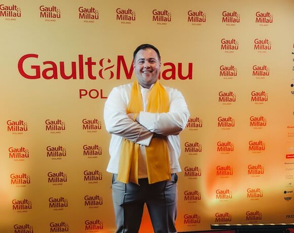 Szef kuchni stoi uśmiechnięty z żółtym szalem i skrzyżowanymi ramionami przed ścianką Gault&Millau Poland w ciepłym świetle.