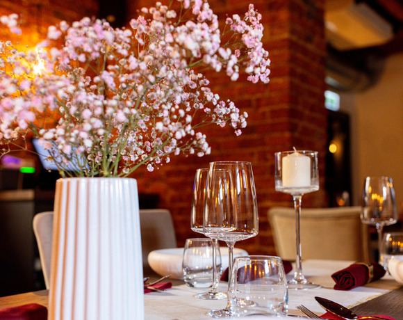 Przytulny stolik w restauracji z białym wazonem z różową gipsówką, kieliszkami, świecą i bordowymi serwetkami przy ceglanej ścianie.