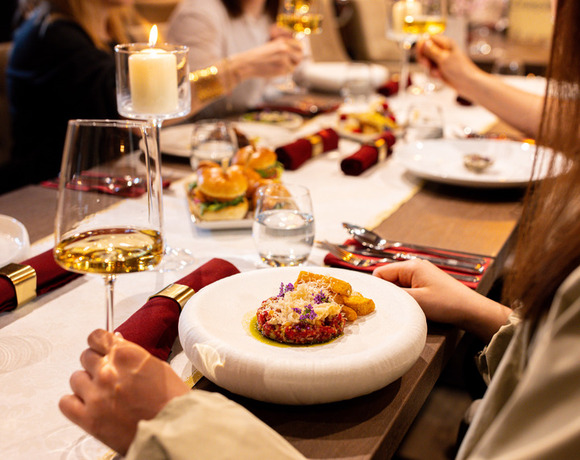 Goście wznoszą kieliszki białego wina przy świecach w restauracji hotelowej, talerz wykwintnego dania z jadalnymi kwiatami i pieczywem.