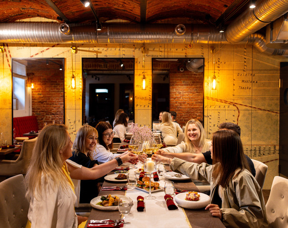 Przyjaciele stukają kieliszkami białego wina przy długim stole w przytulnej restauracji industrialnej z ceglanym murem.