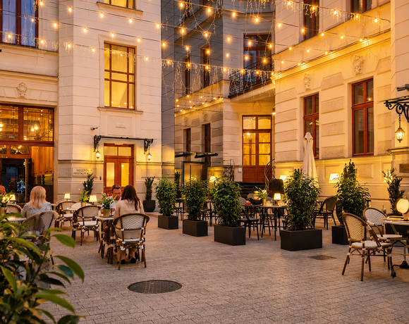 Eleganckie hotelowe patio z restauracją, bistro‑stołami, donicami z roślinami i girlandami świateł, goście rozmawiają i piją.