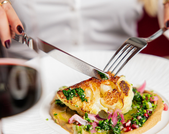 Guest slicing roasted cauliflower with herbs, pickled onions and puree on a white plate in a fine‑dining setting