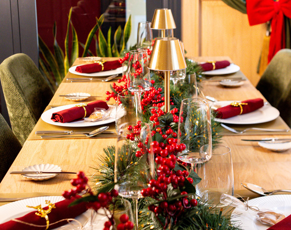 Festive dining table with evergreen garland, red berries, gold lamps, rolled red napkins and elegant place settings.