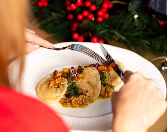Guest cutting stuffed dumplings with fork and knife at a festive table accented by pine greenery, red berries and glasses of red wine.