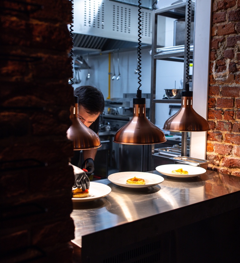 Chef preparing dishes under heat lamps on a stainless steel counter in a professional kitchen with exposed brick wall.
