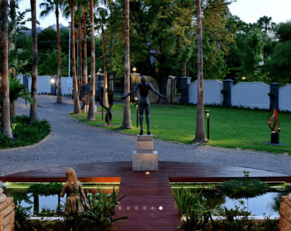 Colonnaded hotel entrance with cobblestone drive, palm-lined path, modern sculptures, pond and wooden bridge lit by evening garden lights.