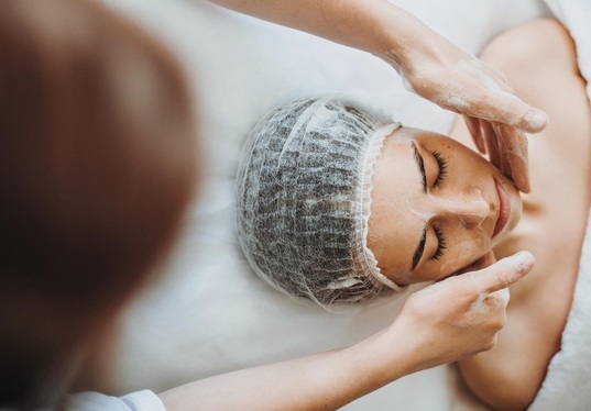 Therapist applies a creamy face mask to a relaxed client in a disposable cap on a spa bed.