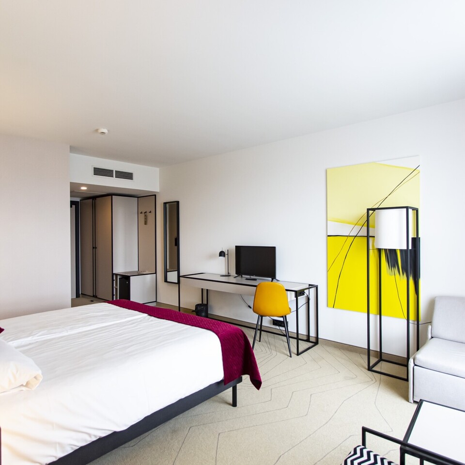 Nowoczesny pokój hotelowy z dużym łóżkiem, biurkiem z komputerem, żółtym krzesłem i miękką sofą przy oknie.