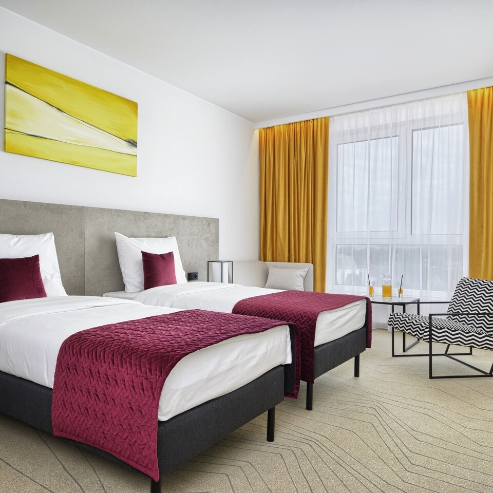 Hotel room with two beds featuring burgundy throws and pillows, yellow curtains, modern armchair, and a sofa by the window.
