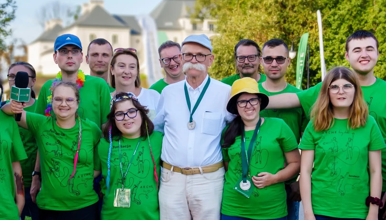 Grupa uczestników w zielonych koszulkach na zewnątrz, starszy mężczyzna w białej koszuli z medalem stoi pośrodku