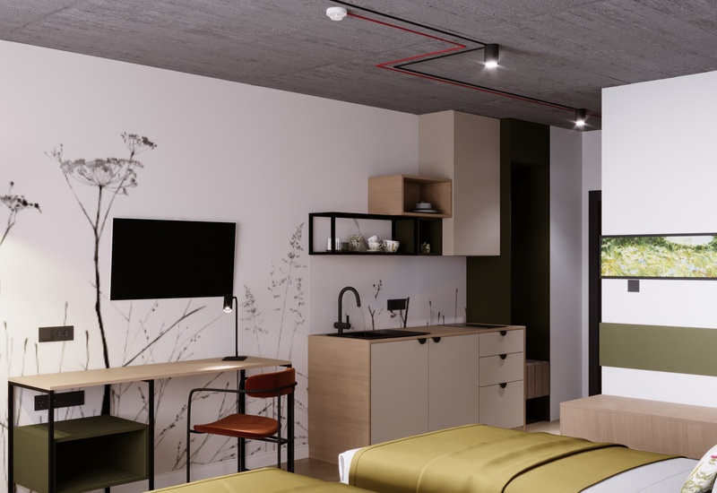 Nowoczesny pokój hotelowy z aneksem kuchennym, biurkiem, telewizorem, zielonymi narzutami i industrialnym sufitem.