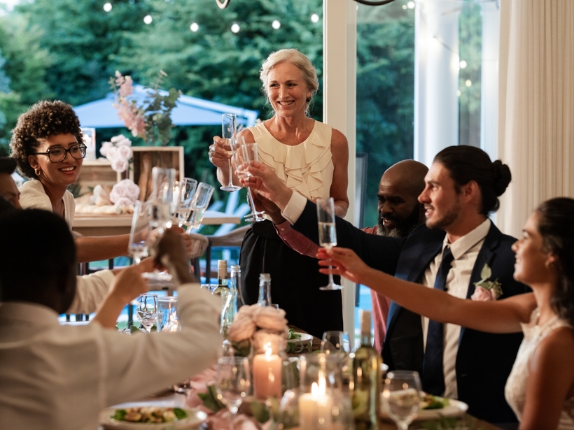 Uroczysty toast przy eleganckim stole z świecami i różowymi kwiatami, goście wznoszą kieliszki na tarasie z widokiem na ogród