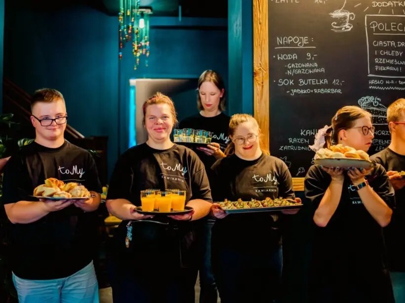 Uśmiechnięty personel kawiarni w przytulnym wnętrzu serwuje soki, wypieki i przekąski na tacach przed tablicą menu.
