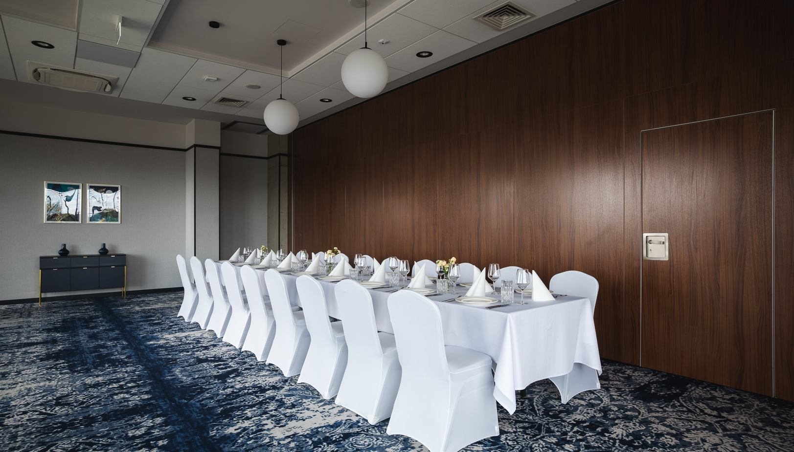 Elegancka sala konferencyjna z długim stołem nakrytym białym obrusem i krzesłami w białych pokrowcach, nowoczesne oświetlenie.