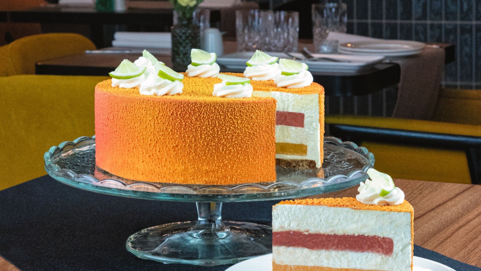 Pomarańczowy tort z kremem i limonką na szklanym stojaku, serwowany w restauracyjnym wnętrzu z żółtymi fotelami.